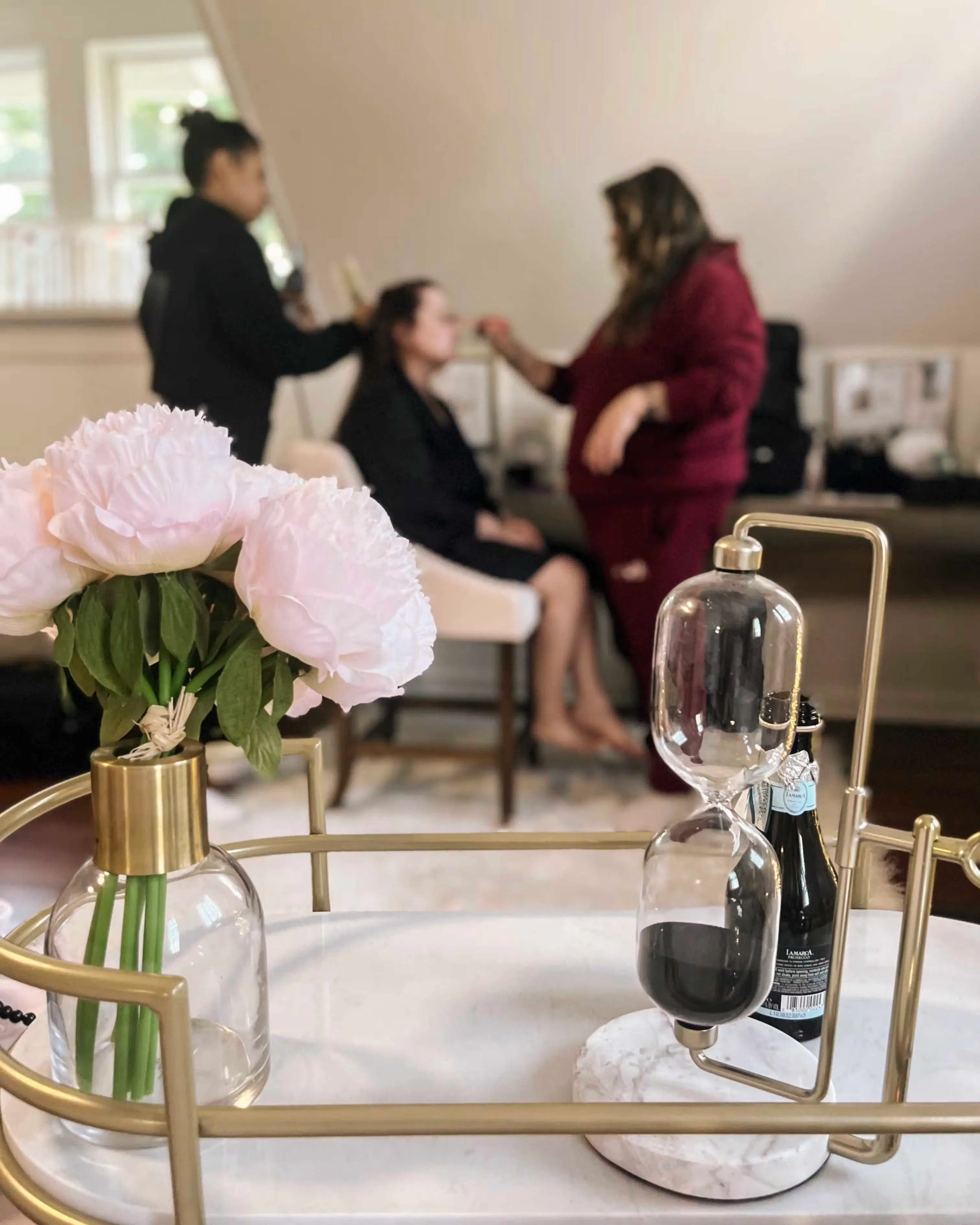 Close-up of a table with a glass vase holding pink flowers and a modern hourglass with black sand, with two people in the background applying makeup to a seated woman.