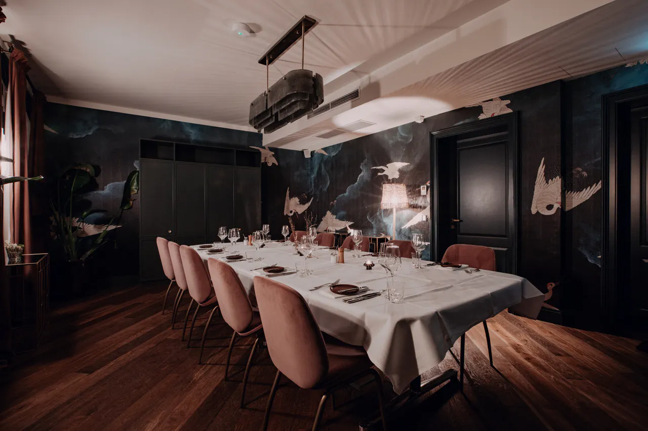 Esszimmer mit langem gedecktem Tisch, rosa gepolsterten Stühlen, dunklen Wänden mit Vogelmotiven und Holzboden. Deckenbeleuchtung und Stehlampe sorgen für warmes Licht.