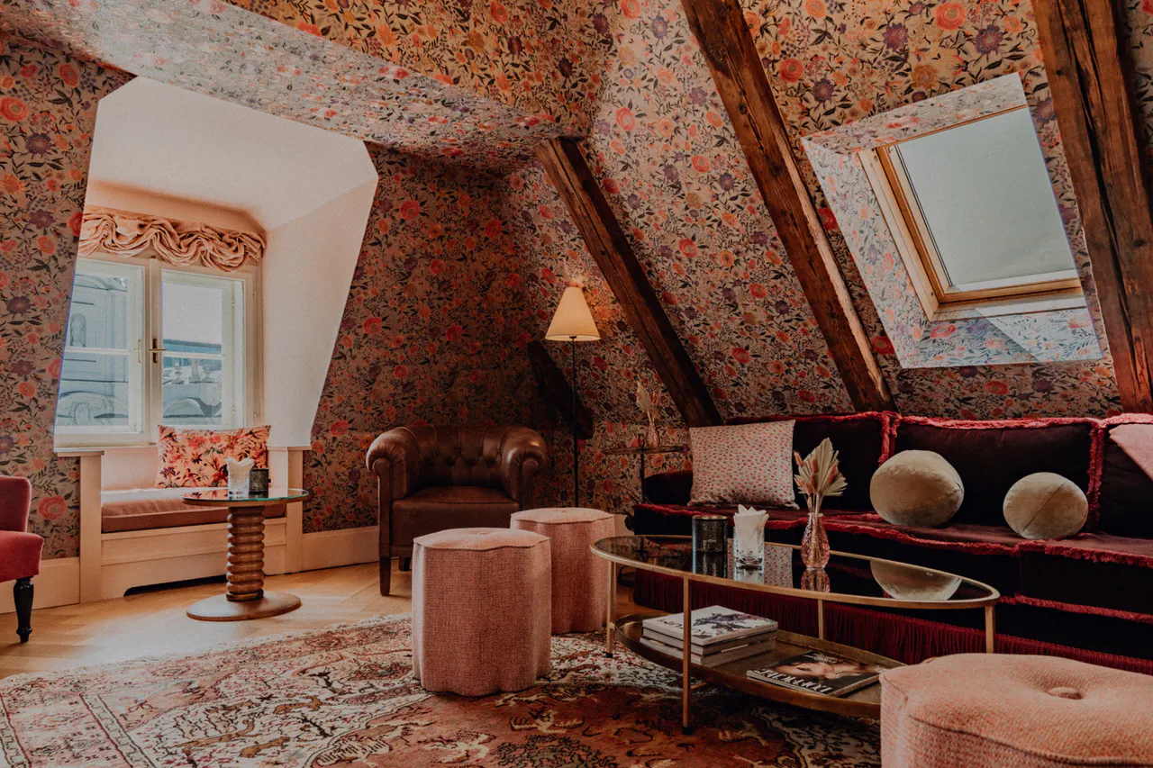 Gemütliches Wohnzimmer unterm Dach mit floralem Tapetenmuster, dunklem Samtsofa, Ledersessel, zwei Poufs, großem Teppich und einem Fenster mit Sitzbank.