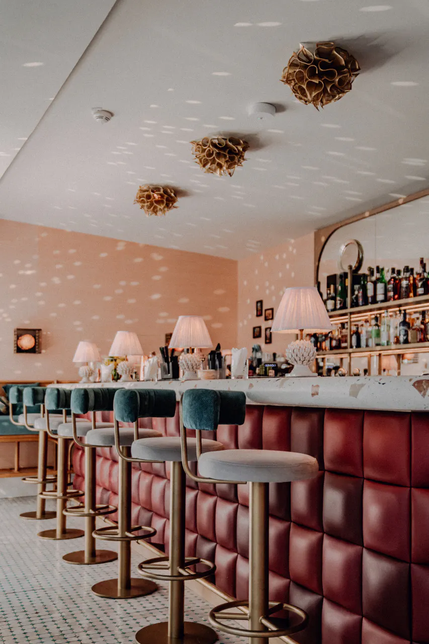 Bar mit rot gepolstertem Tresen, grünen Barhockern, dekorativen Lampen auf der Theke und Regalen mit Flaschen im Hintergrund.