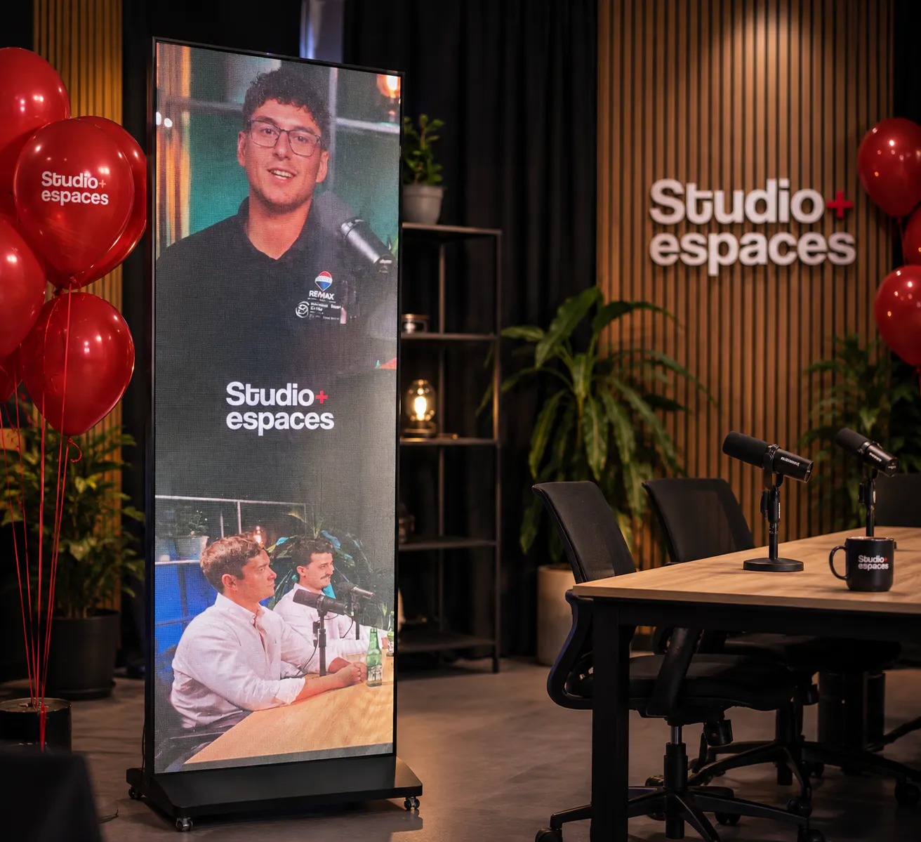 Digital screen showing two men speaking into microphones, three red balloons with "Studio+ espaces," a table with microphones and a mug, and a wall with "Studio+ espaces" sign.