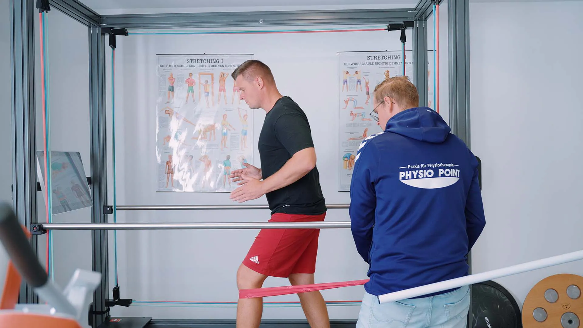 Mann trainiert mit Theraband um die Beine, betreut von Physiotherapeut in Sportpraxis mit Trainingsgeräten und Stretching-Postern im Hintergrund.