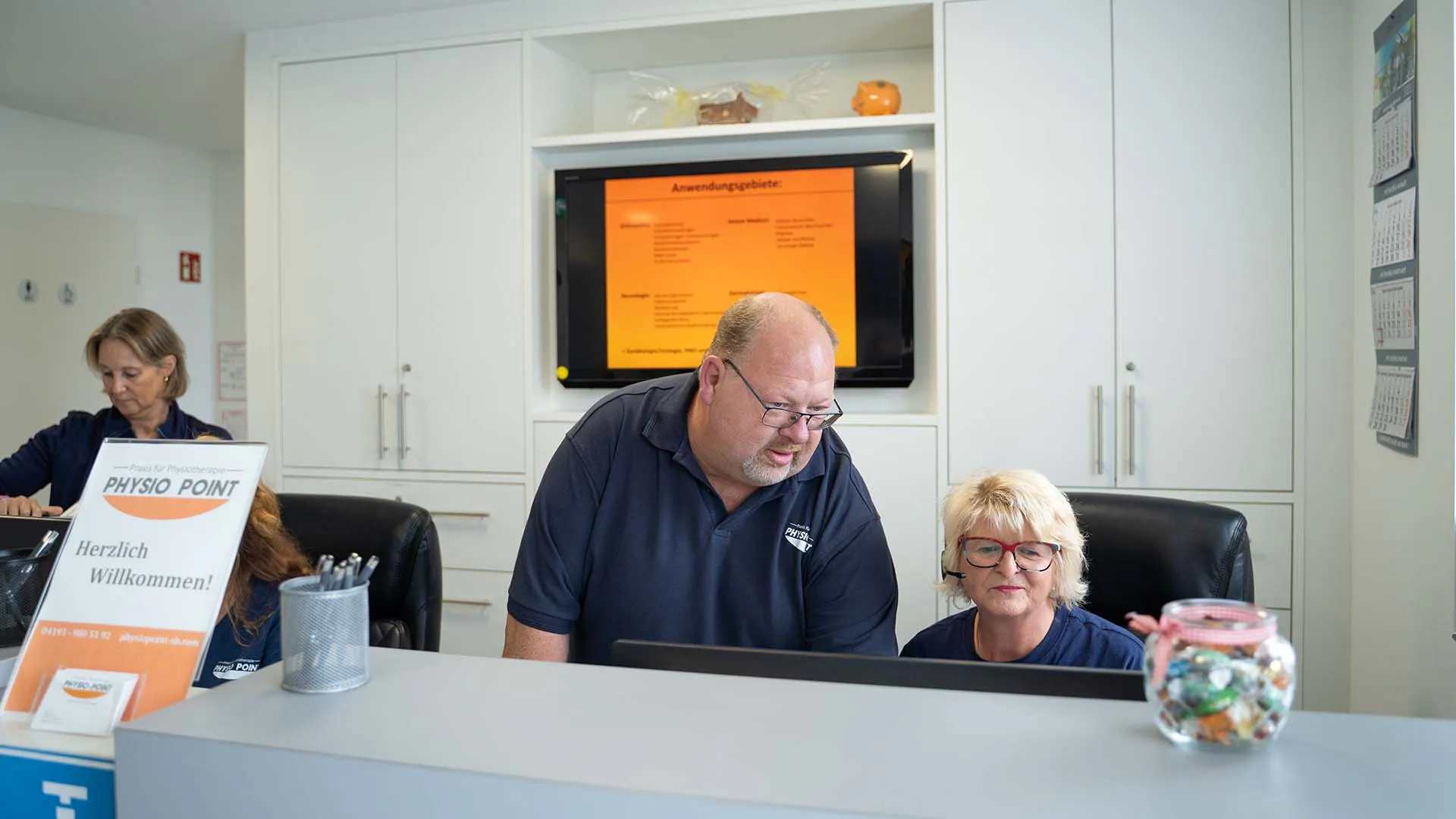 Zwei Personen an einem Empfangstresen in einer Physiotherapiepraxis, eine Frau sitzend mit Headset und ein Mann stehend, dahinter weiße Schränke und ein Bildschirm mit Text.
