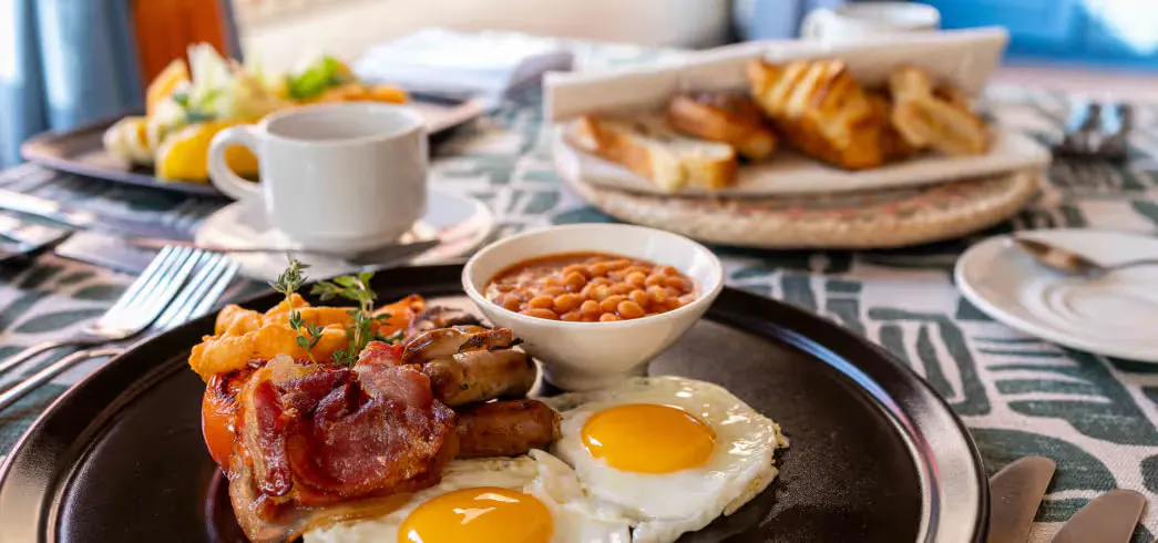 Two fried eggs, bacon, sausage, grilled tomato, baked beans, and hash browns on a black plate with a cup of coffee and croissants in the background.