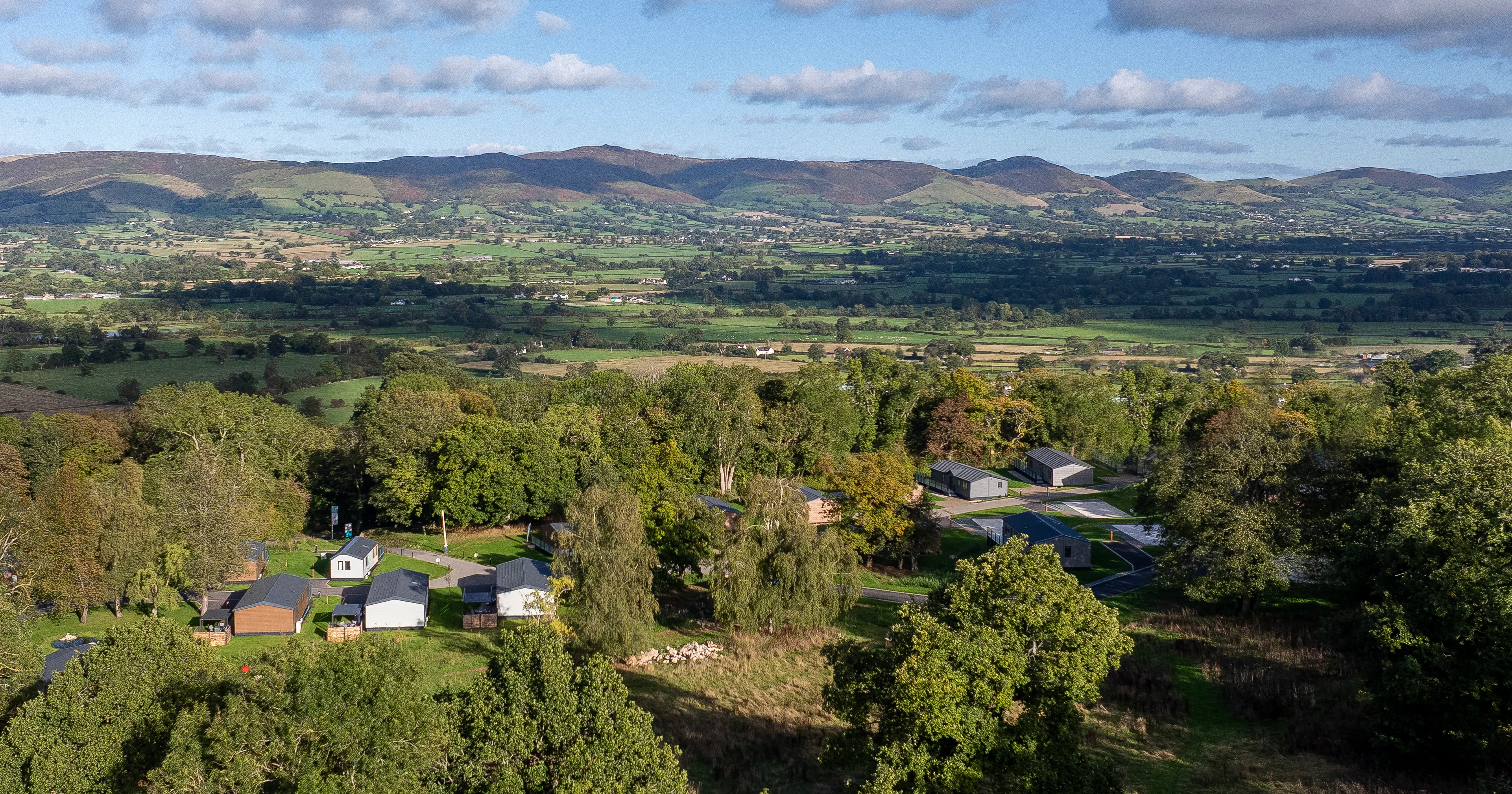 Rural landscape with scattered houses surrounded by trees, green fields, and distant hills under a partly cloudy sky.