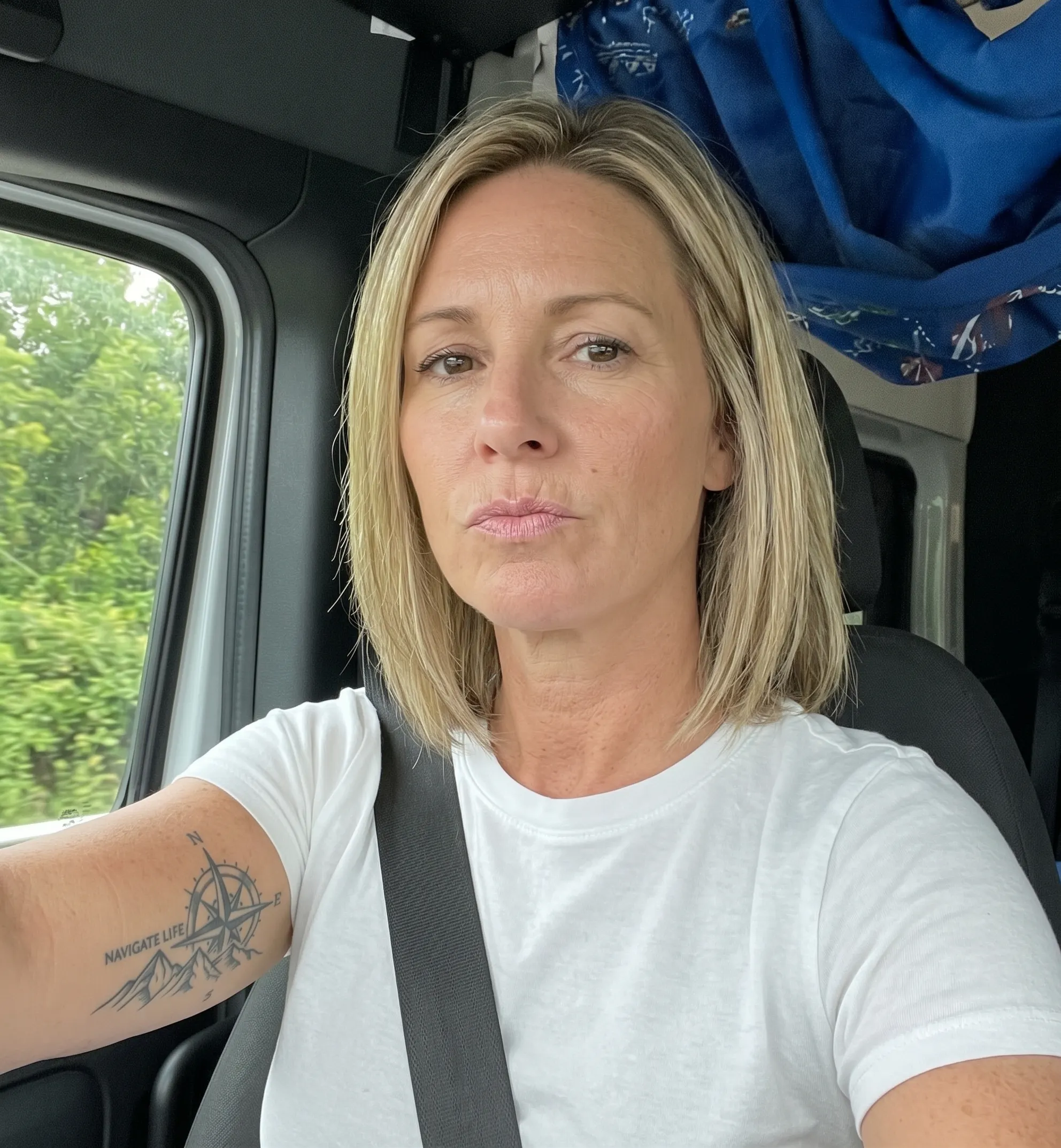 Woman with shoulder-length blonde hair wearing a white t-shirt and black seatbelt, sitting in a vehicle driver's seat, with a compass and mountain tattoo on her left arm.