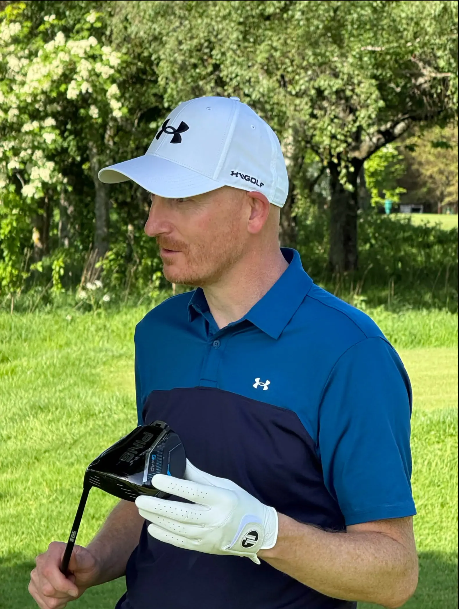 Man wearing a white Under Armour cap, blue golf shirt, and white golf glove holding a Ping golf club on a grassy area with trees in the background.