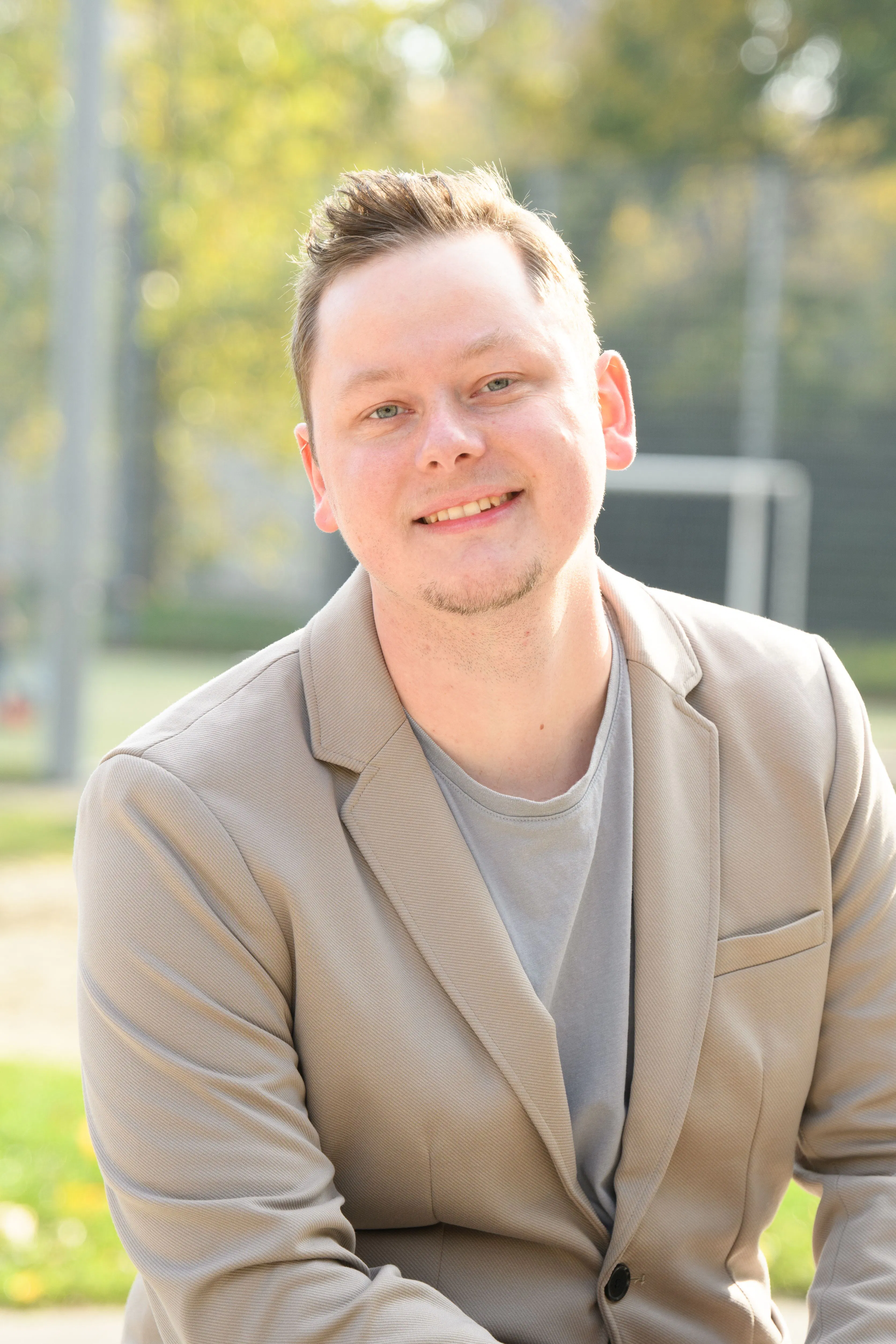 Mann mit kurzem braunem Haar trägt hellbeige Sakko und graues T-Shirt, lächelt in die Kamera, unscharfer grüner Hintergrund mit Bäumen und Fußballtor.