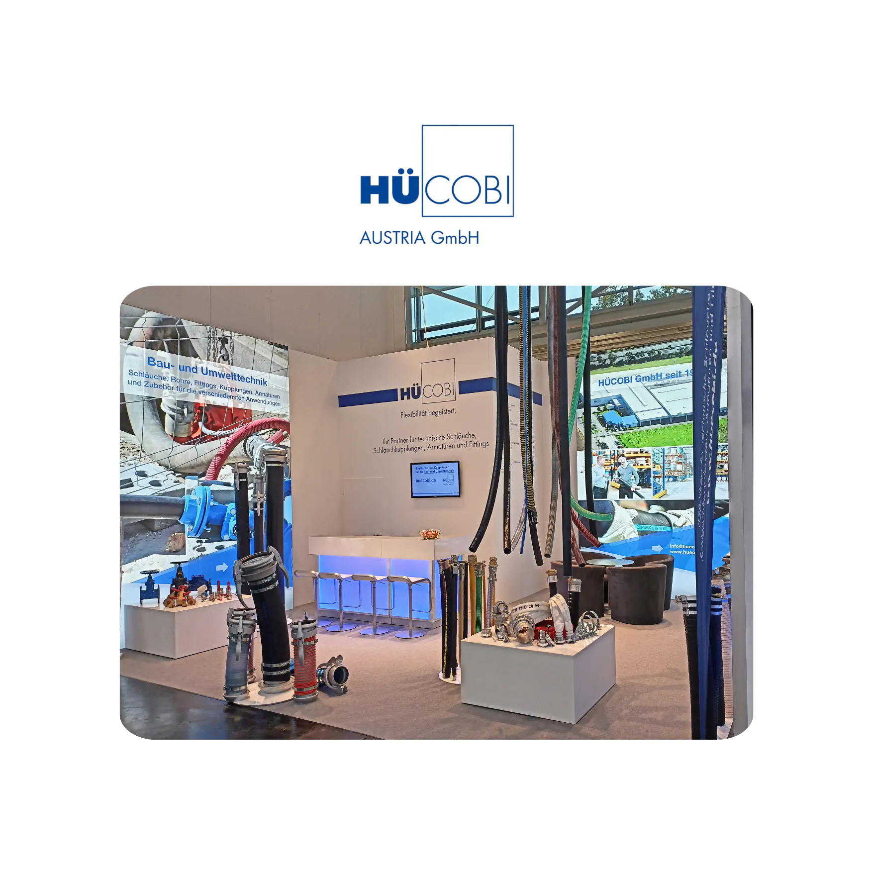 Messestand der Firma HÜCOBI Austria GmbH mit technischen Schläuchen, Kupplungen, Armaturen und Fittings, beleuchtetem Tresen und Informationsbildschirmen.