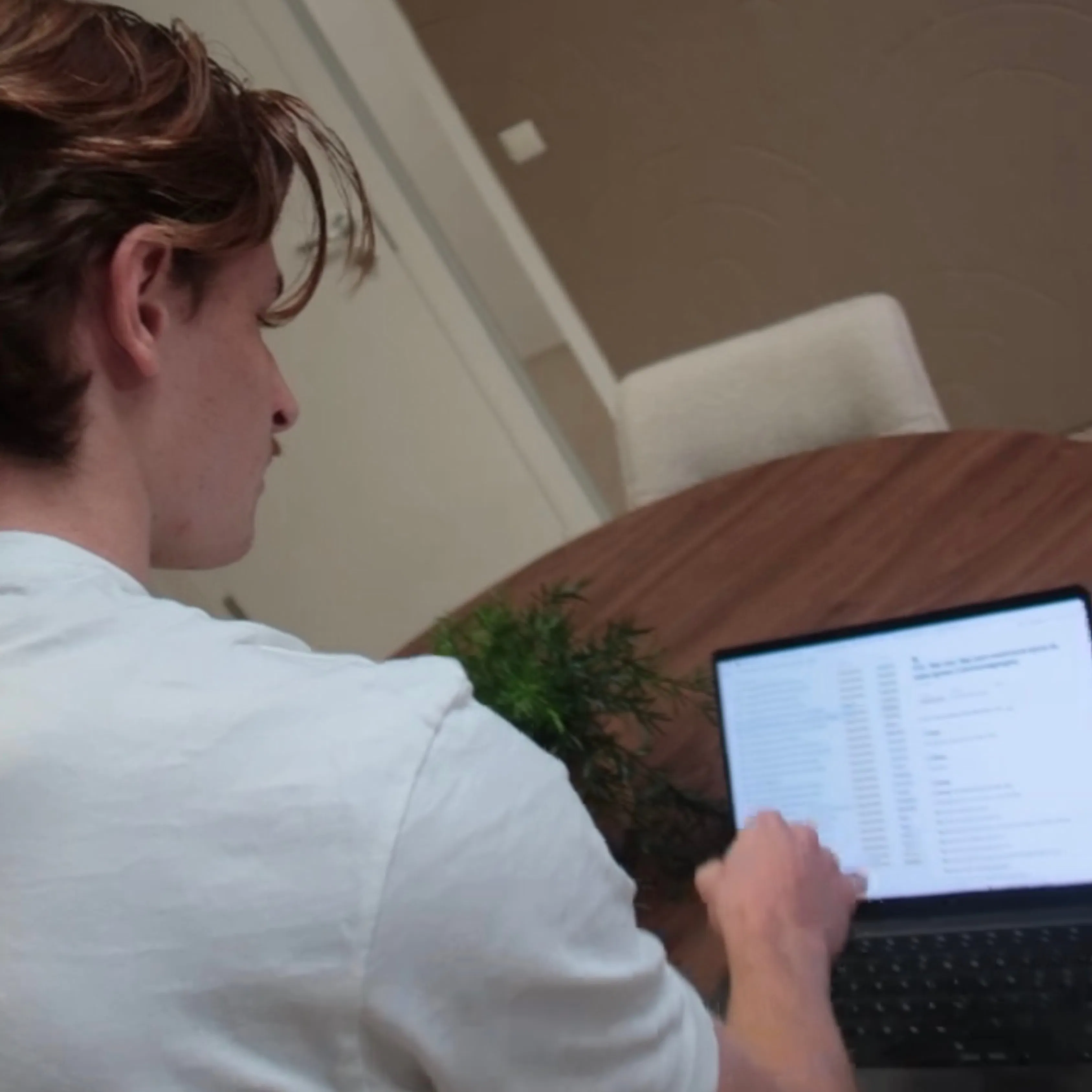 Person in a white shirt working on a laptop at a round wooden table with a small green plant, viewed from behind.