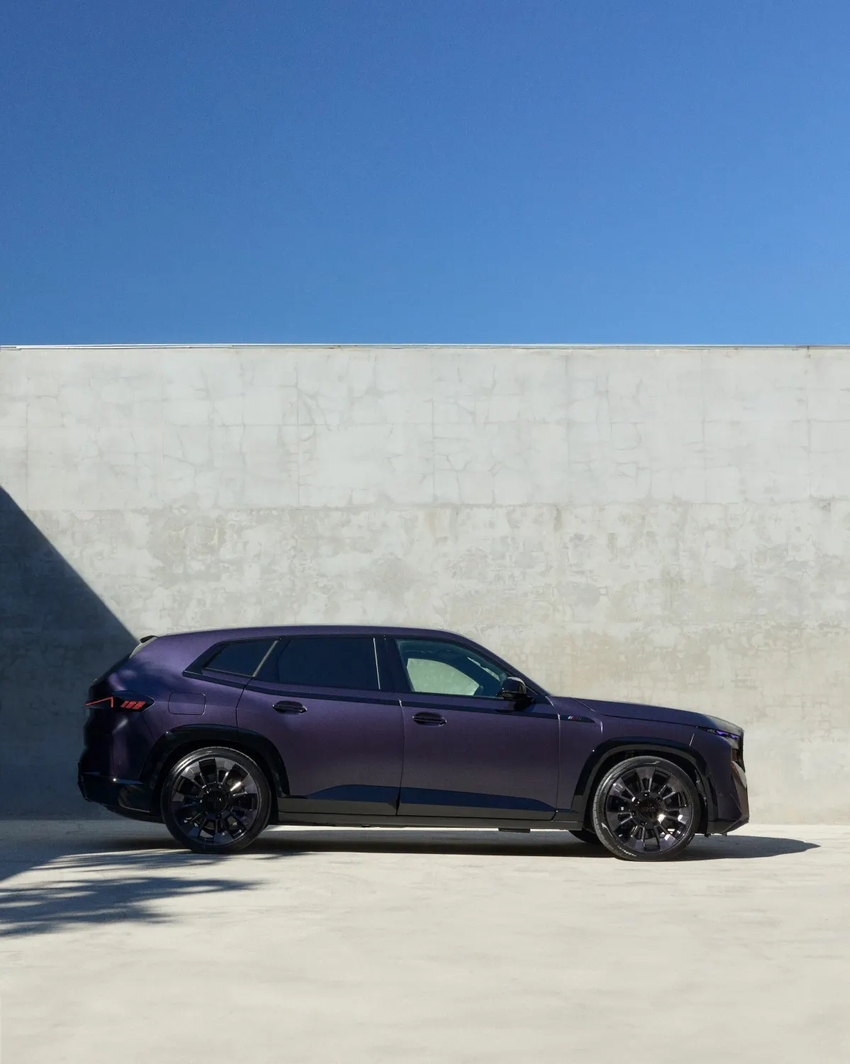 Side view of a dark purple modern SUV parked next to a light grey concrete wall under a clear blue sky.