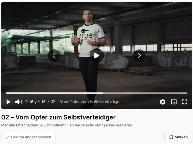 Mann in hellem T-Shirt mit der Aufschrift „True Martial Arts“ steht in einer großen, leeren Halle und spricht vor einer Kamera.