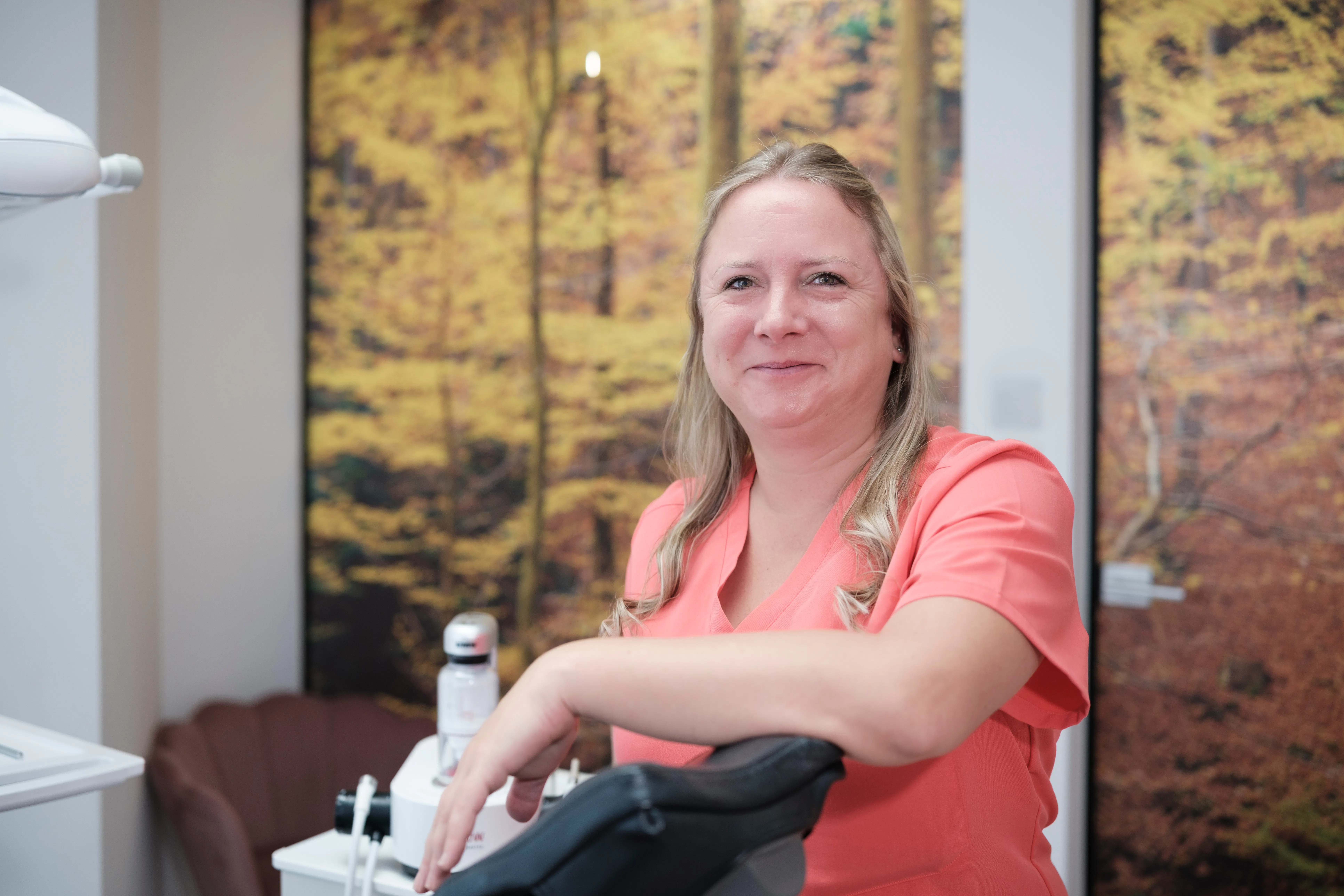 Frau in orangener Arbeitskleidung lehnt lächelnd an einem Zahnarztstuhl, im Hintergrund ein Bild mit Herbstwald.