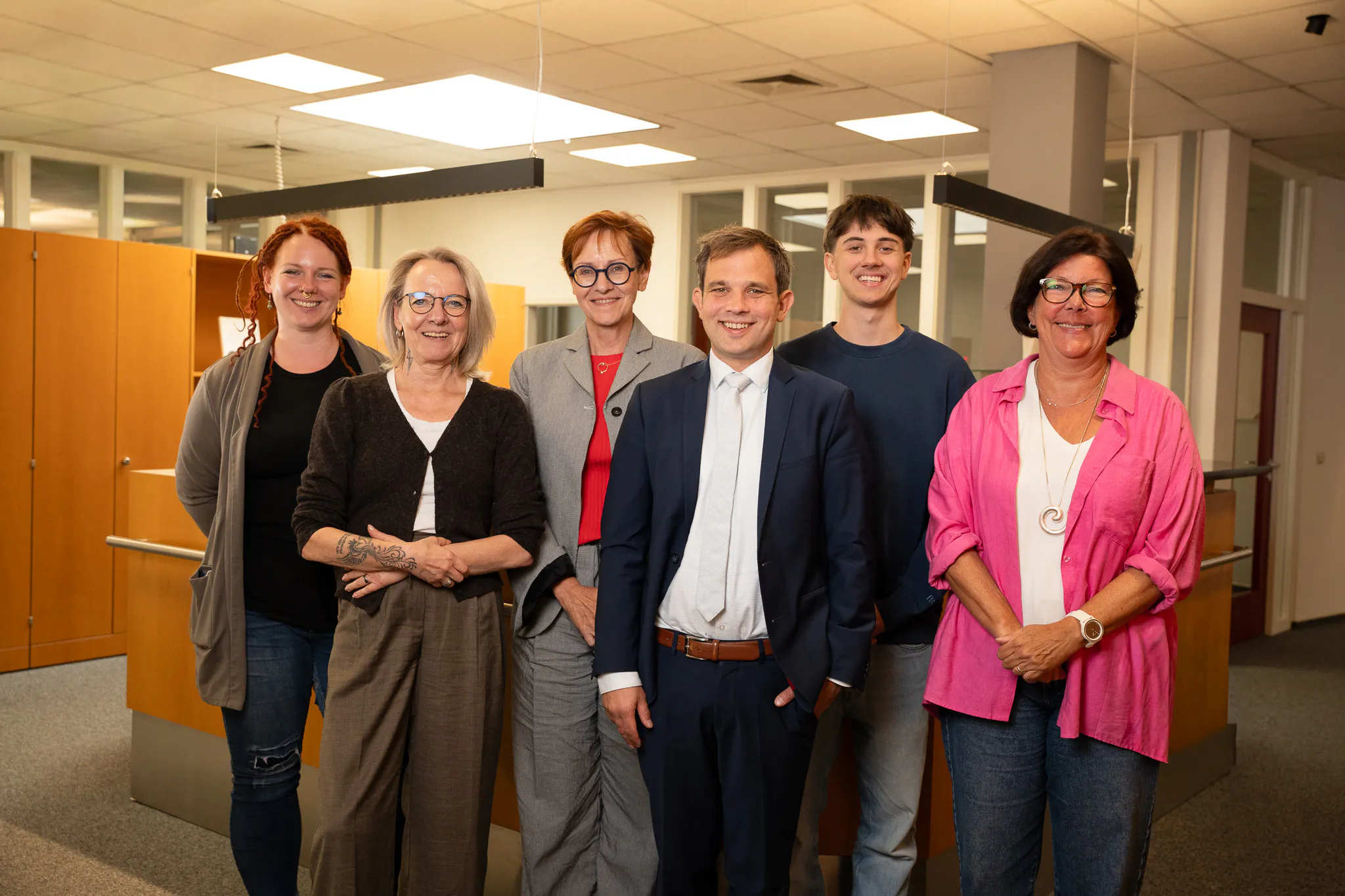 Sechs Personen, drei Frauen und drei Männer, stehen nebeneinander in einem Büro und lächeln in die Kamera.