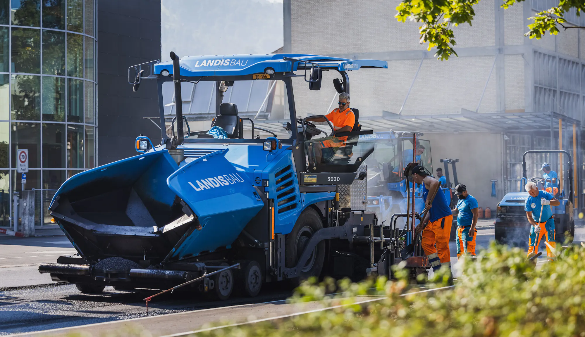 Blauer Landis-Bau-Belagsfertiger mit Maschinist im Einsatz, daneben das Team beim Belagseinbau