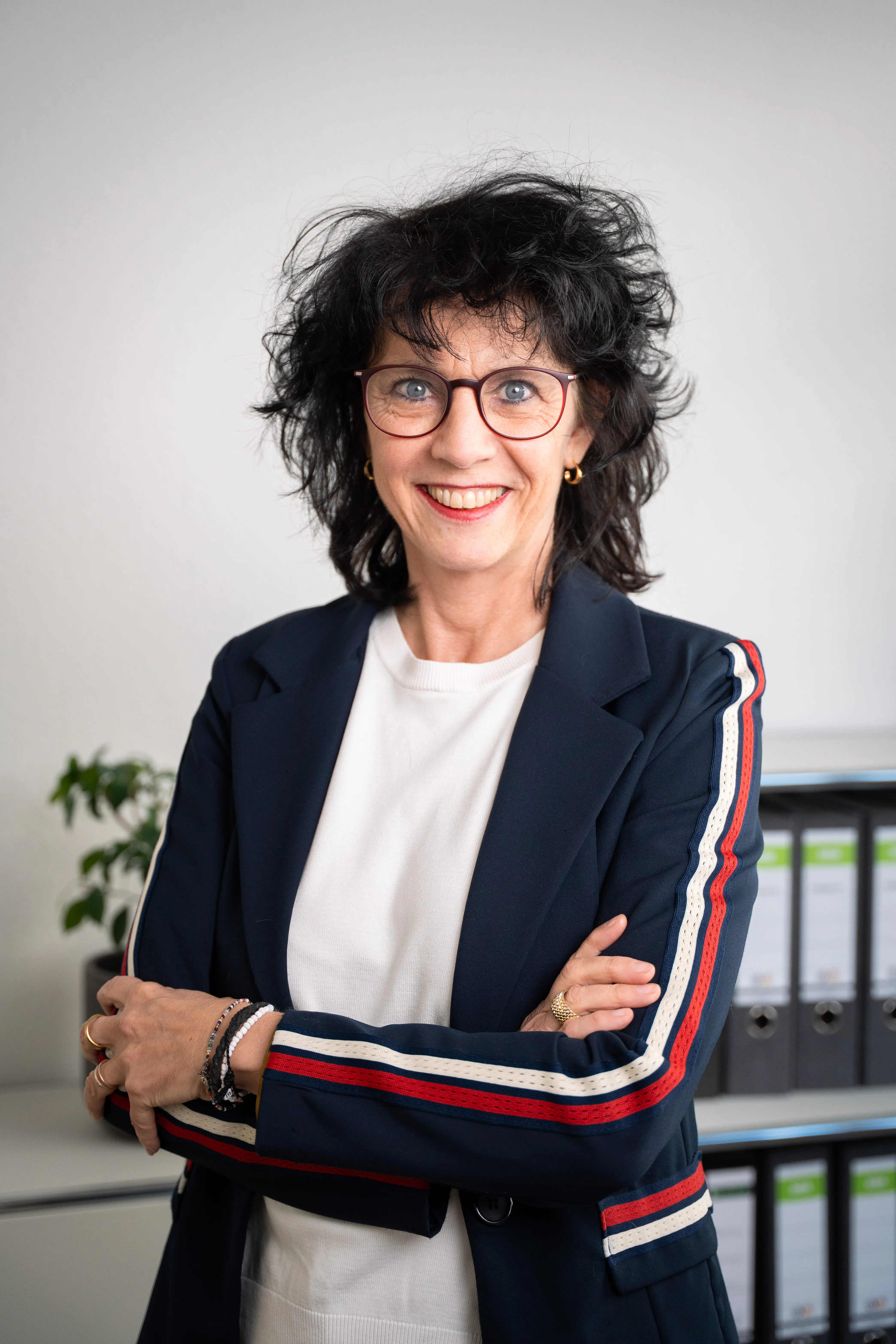 Frau mit schwarzen lockigen Haaren, Brille und blauem Blazer mit rot-weißen Streifen auf den Ärmeln, lächelt mit verschränkten Armen vor einem Regal mit Ordnern.