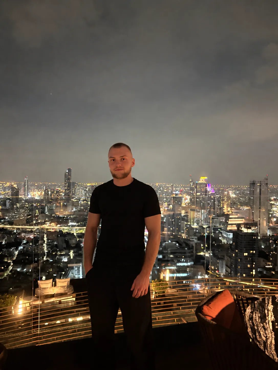 Portrait of David Andreju on a rooftop at night, the lit city skyline behind him