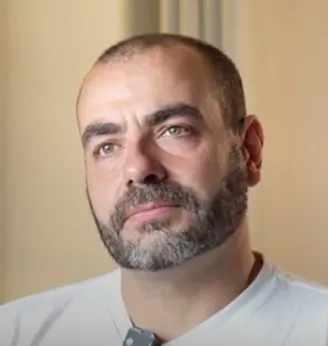Man with short dark hair and a beard, wearing a light-colored shirt, looking slightly to the side against a plain background.