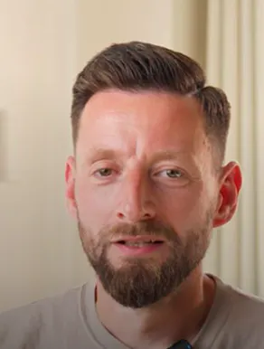 Close-up of a man with short brown hair and a beard, looking slightly downward with a neutral expression, against a blurred light-colored background.