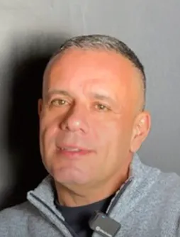 Man with short gray hair and light skin wearing a gray zip-up jacket and a black undershirt against a plain gray background.