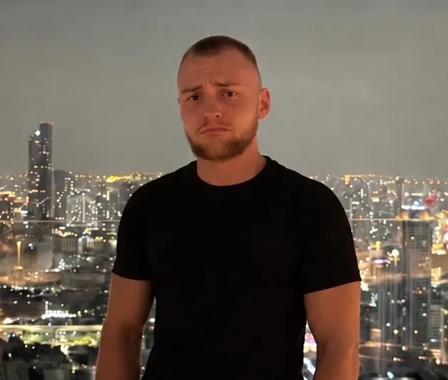 Young man with short hair and beard wearing a black t-shirt standing against a cityscape with numerous illuminated buildings at night.