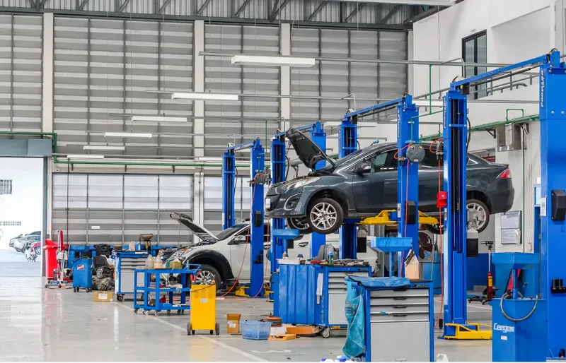 Car repair workshop with multiple hydraulic lifts, two cars raised and open hoods, surrounded by blue tool cabinets and equipment.