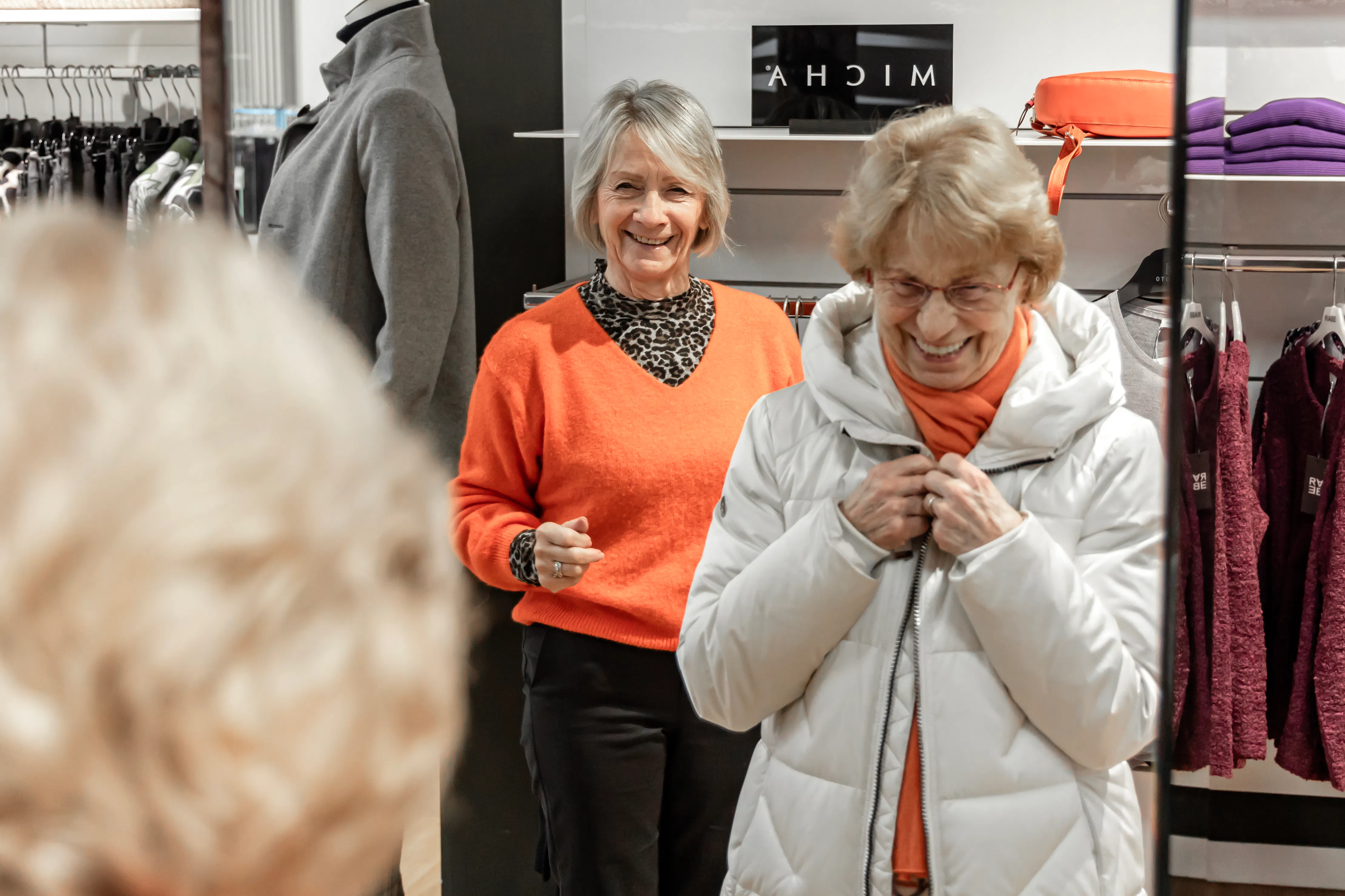Two older women smiling in a clothing store; one wearing an orange sweater and leopard print top, the other adjusting a white puffer jacket with an orange scarf in front of a mirror.