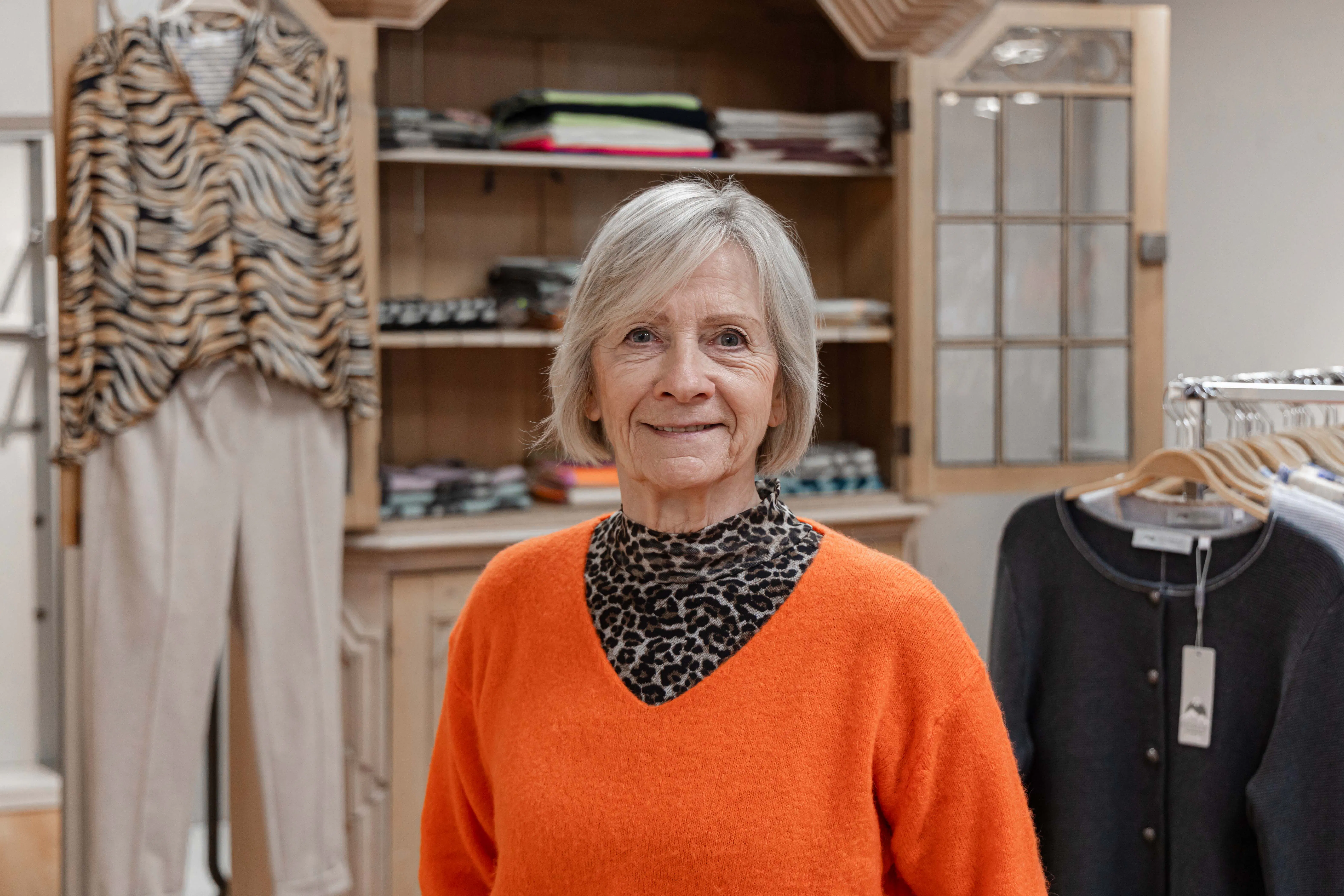 Ältere Frau mit kurzem grauem Haar, trägt orangefarbenen Pullover und Leopardenmuster-Oberteil, steht vor einem Kleiderschrank und Kleiderbügel mit Kleidung.