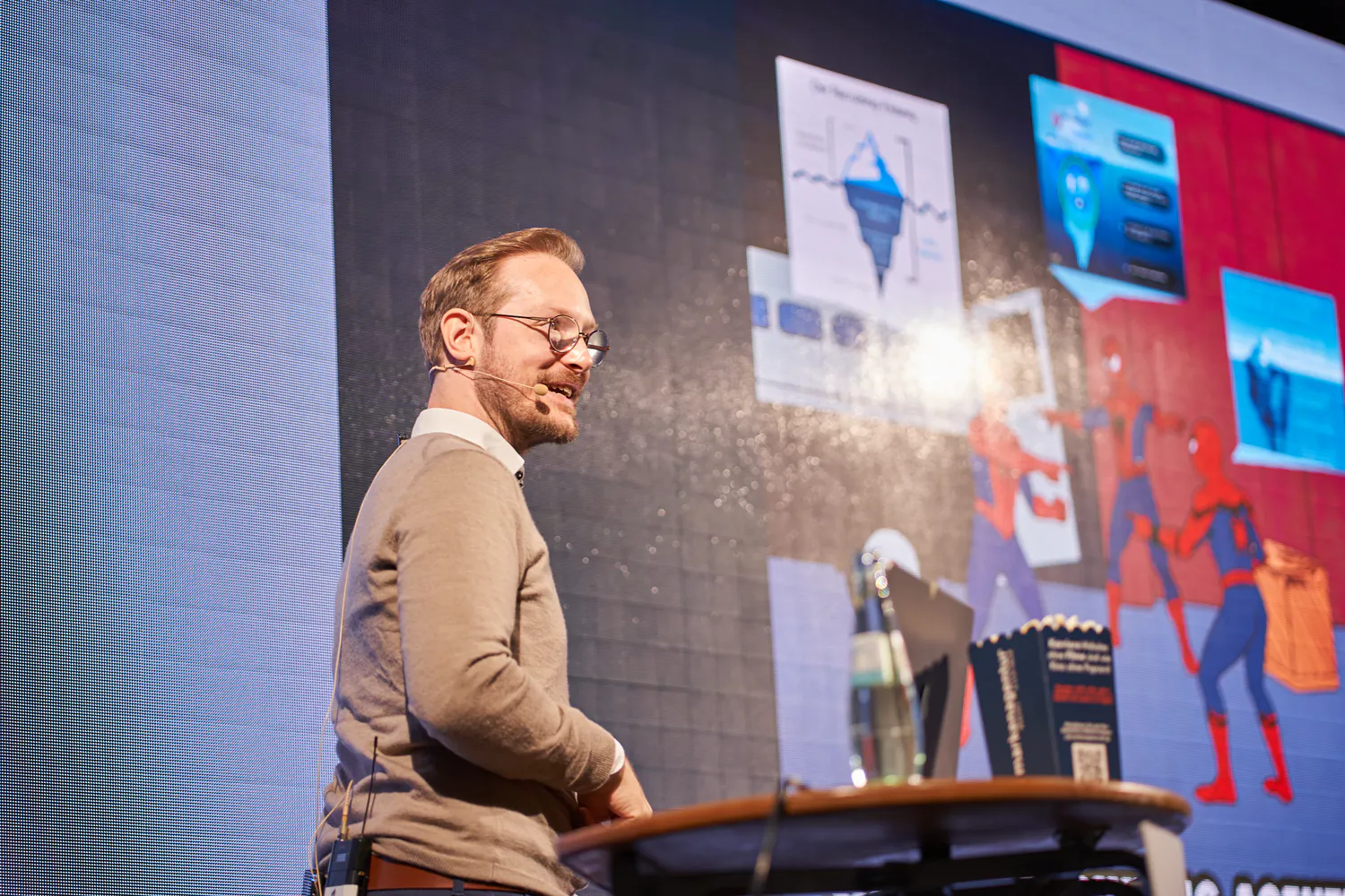 Sprecher mit Brille und Headset hält einen Vortrag auf einer Bühne vor einer großen Leinwand mit Eisberg-Grafik.