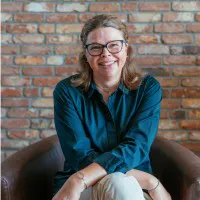 Person with glasses and a blue shirt sitting in a brown chair in front of a brick wall, smiling.